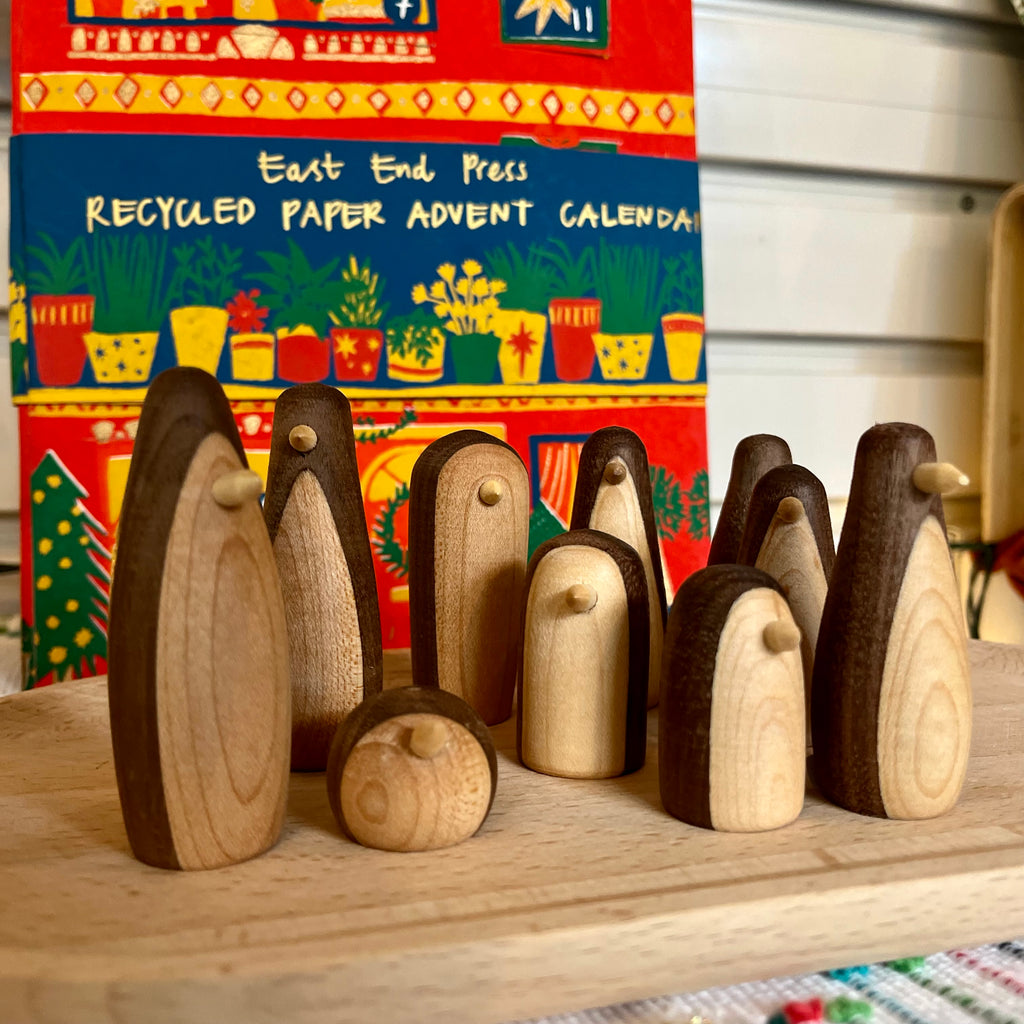 Wooden penguin figures on a wooden surface with a colorful recycled paper advent calendar in the background.