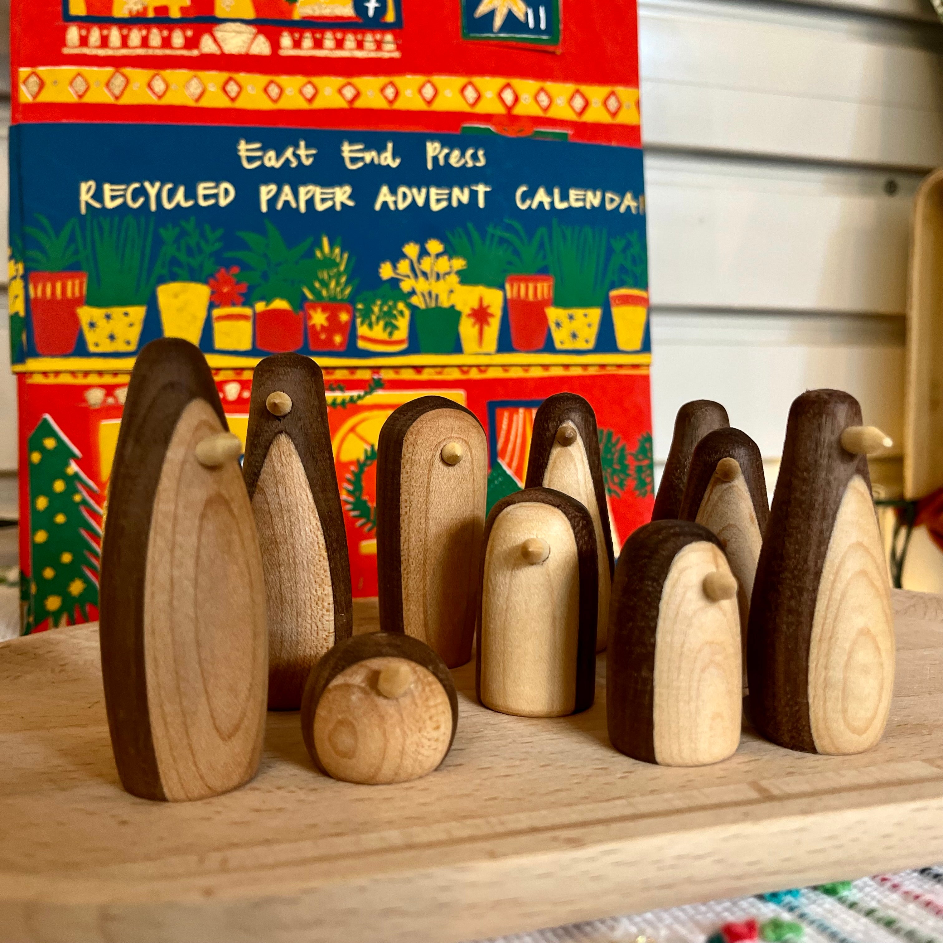 Wooden penguin figures on a wooden surface with a colorful recycled paper advent calendar in the background.