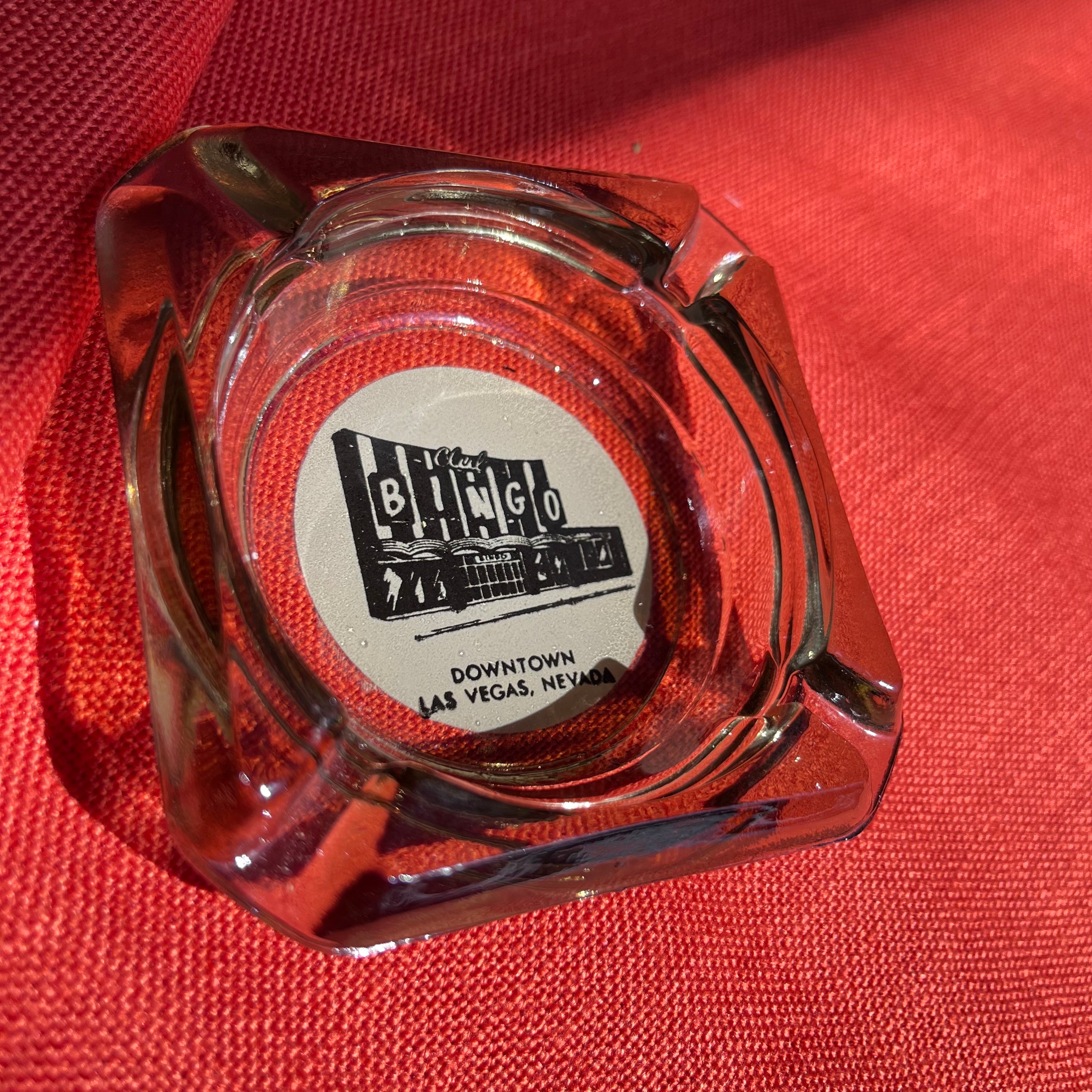 Clear glass ashtray with a red center and 'Club Bingo' logo on a red fabric background