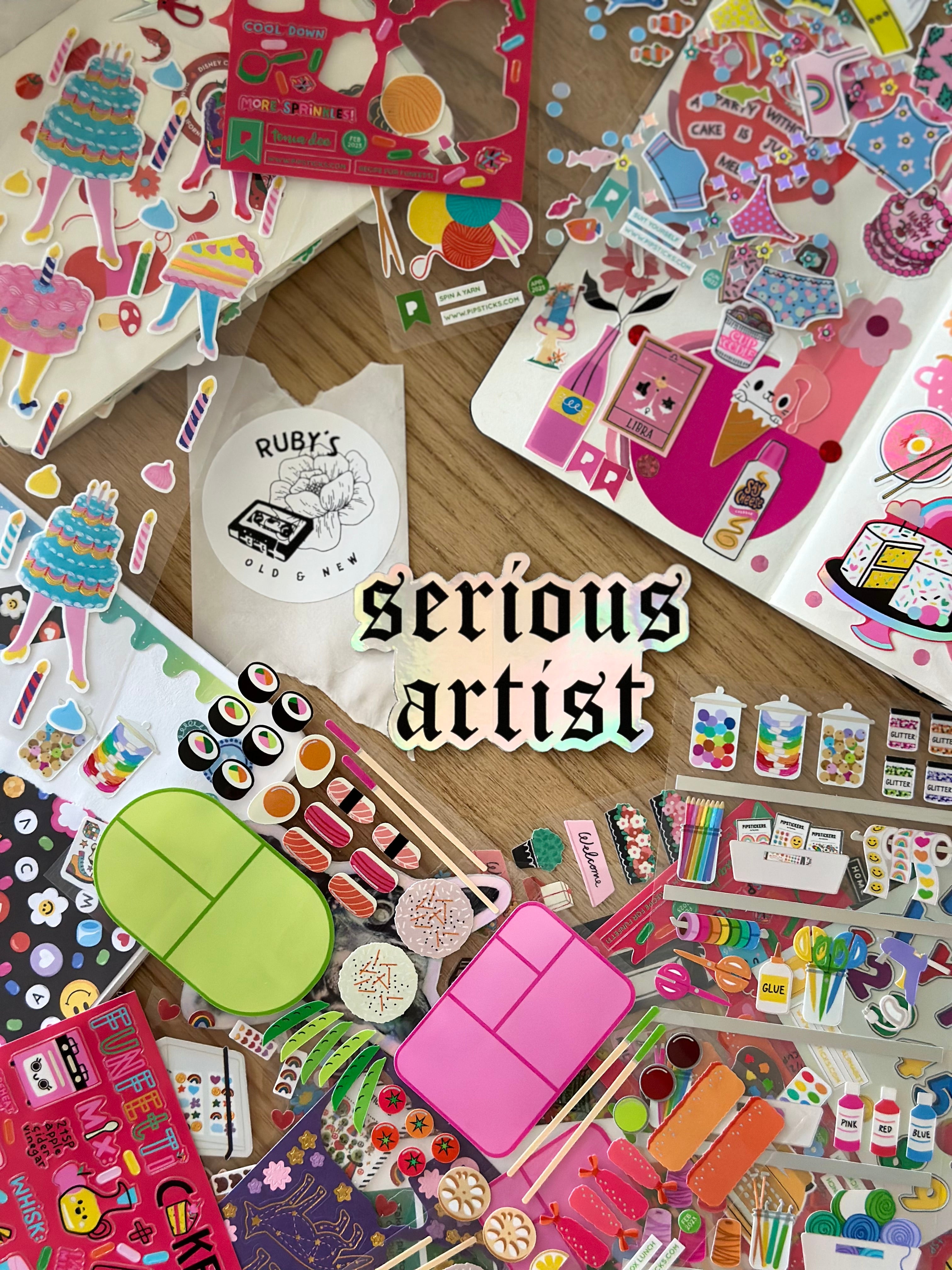 a flatlay of stickers with a holographic sticker in the middle that says "serious artist"