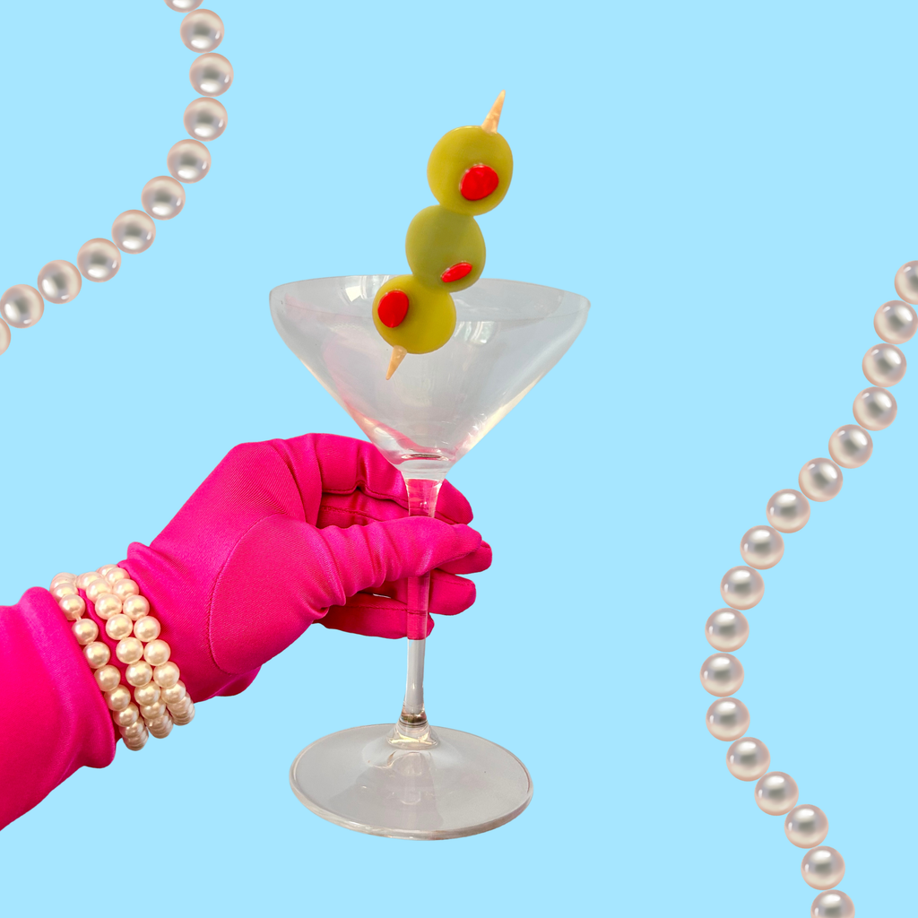 Hand in pink glove holding a martini glass with an olive hair clip inside, pearl necklace on blue background