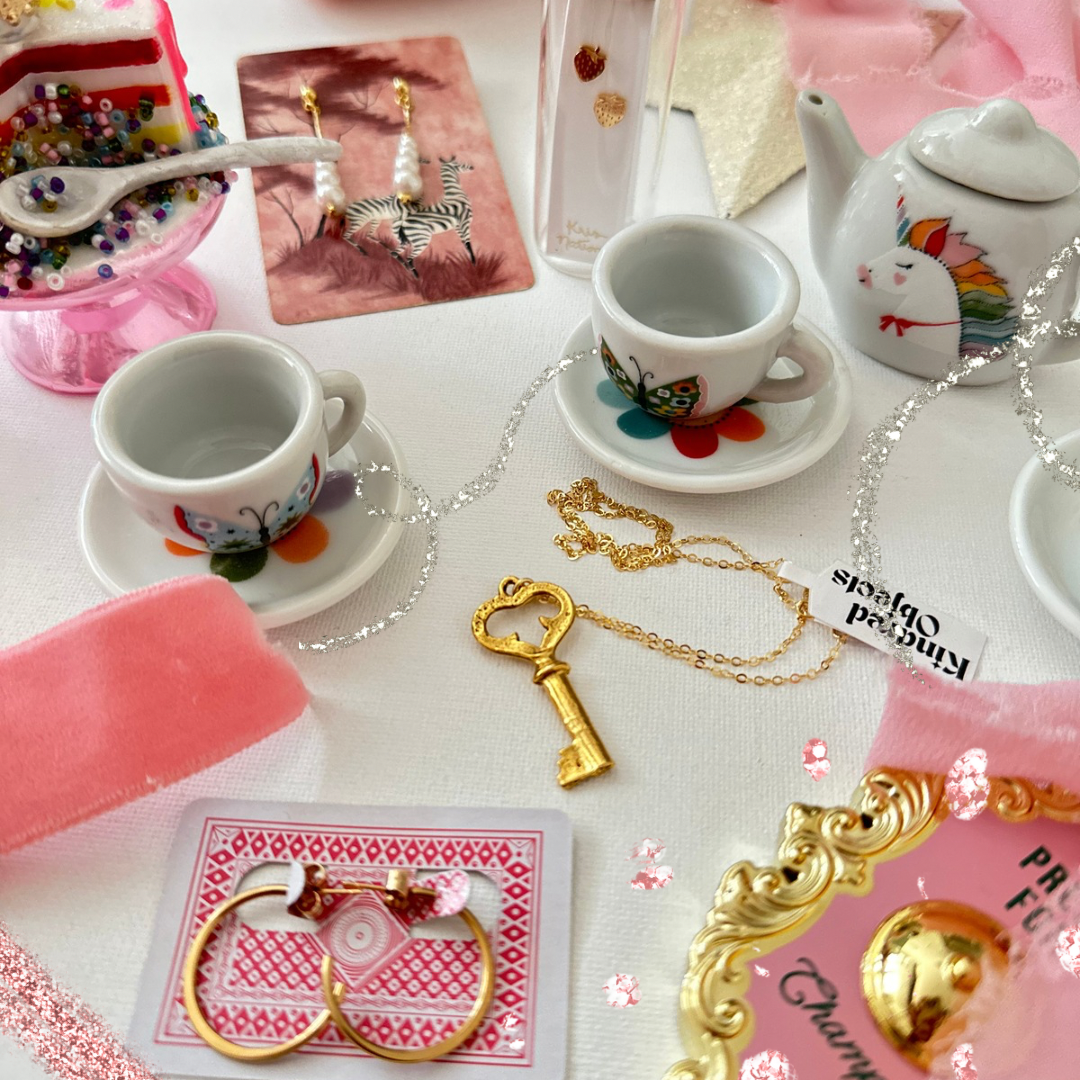 Decorative items including a teapot, cups, key, and jewelry on a white surface with pink accents.