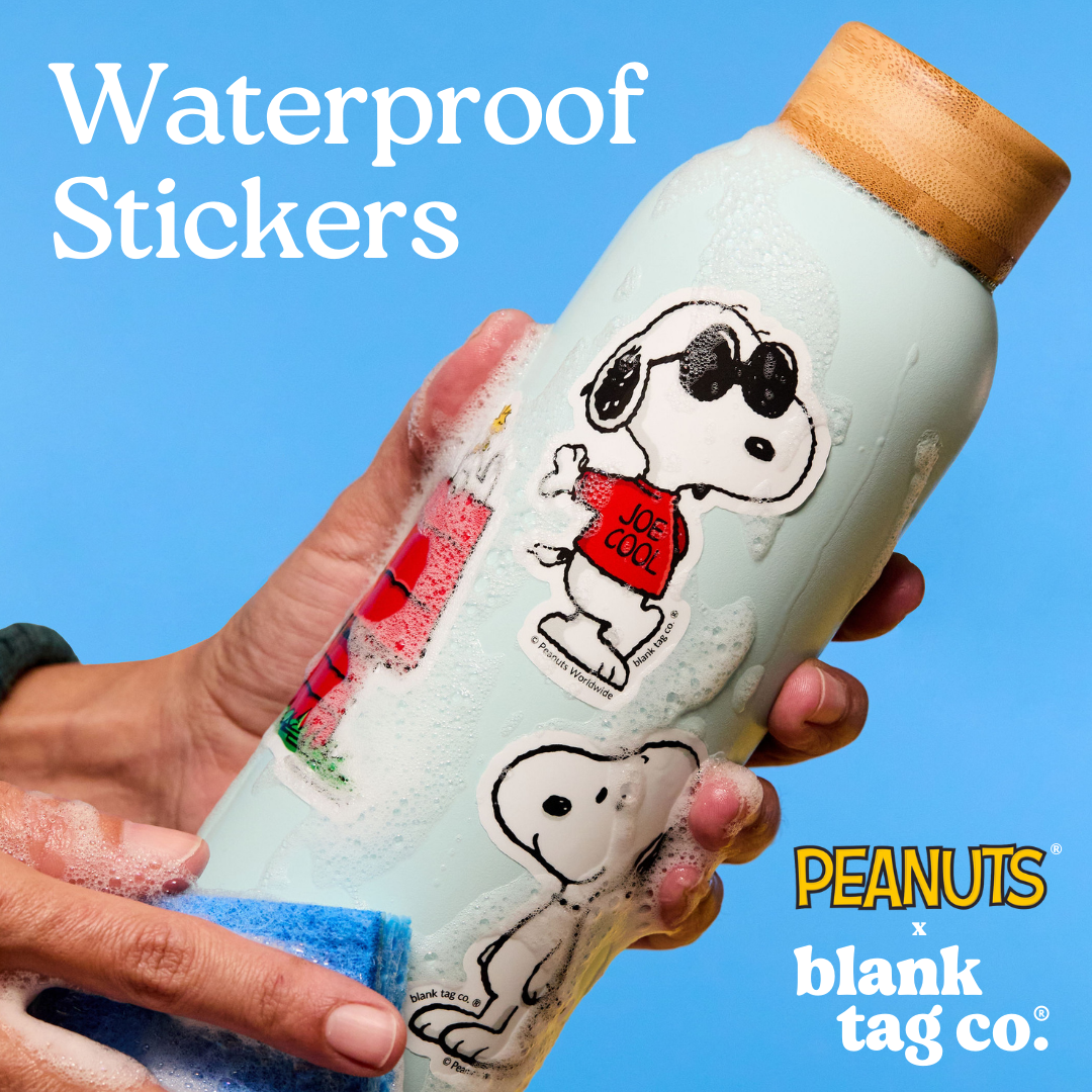 Hand holding a water bottle with Snoopy stickers against a blue background, featuring 'Peanuts' and 'blank tag co.' branding.
