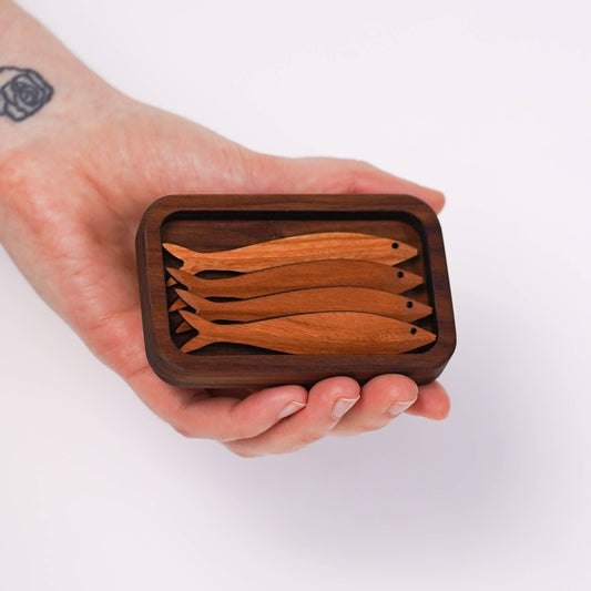 A hand with a tattooed wrist displays a handheld sized wooden replica of tinned fish, the "tin" and "fish" both handcarved from cherry wood.